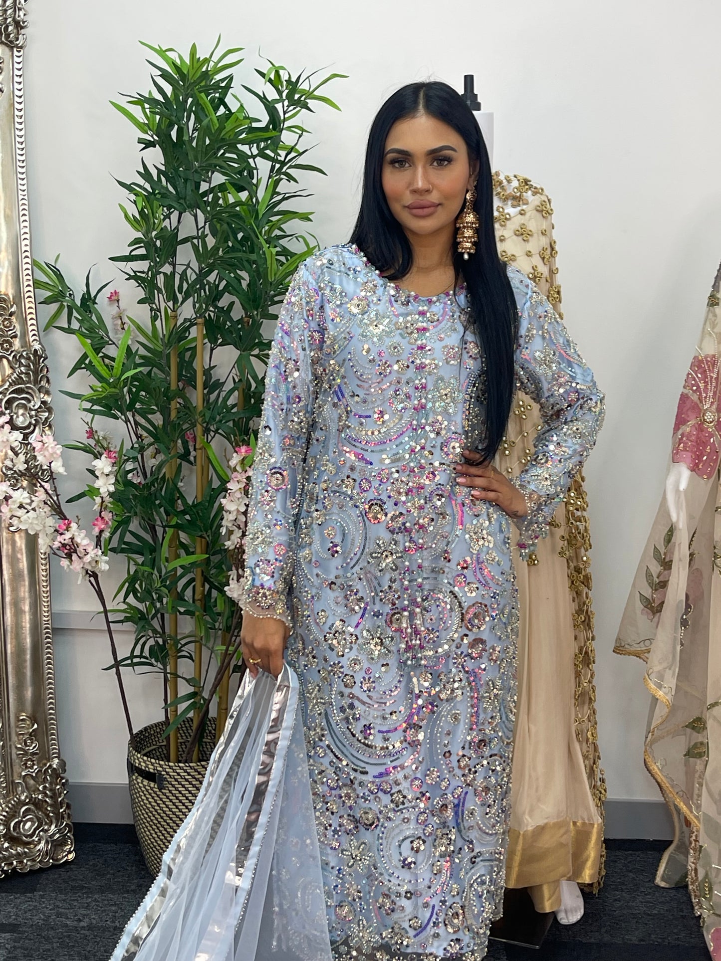 Woman in an ornate dress standing next to decorative mirrors and plants.