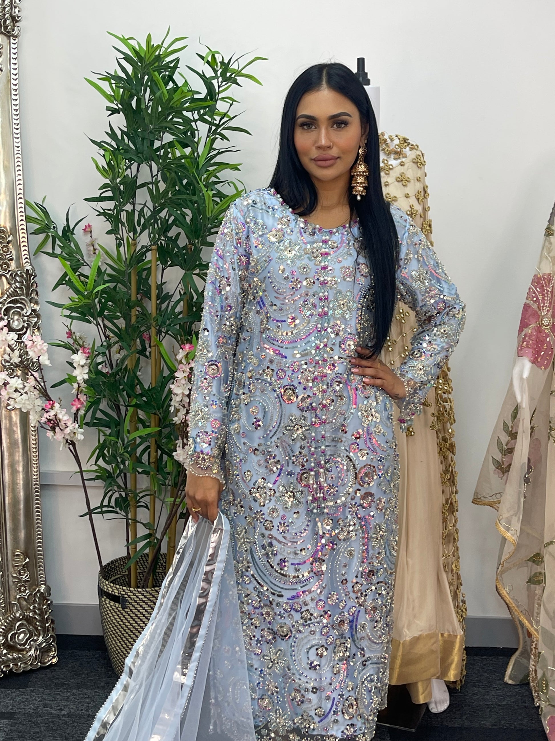 Woman in an ornate dress standing next to decorative mirrors and plants.